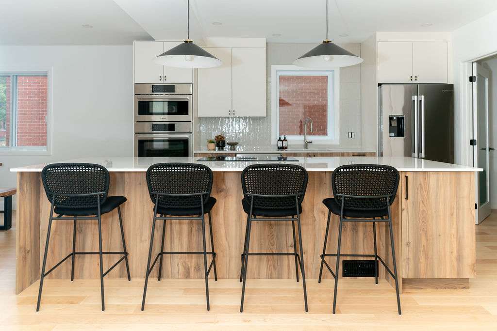 Cuisine fini blanche et bois. Four encastré au mur. Réfrigérateur sur le mur du fond. Une fenêtre se trouve au-dessus de l'évier. Dosseret de céramique installé à la vertical blanc lustré. Armoires blanches mates. Gros îlot entièrement en bois. Rangement intégrés avec quatre places assises. Tabouret noirs avec dossier. Comptoir en quartz blanc. Plaque de cuisson intégrée dans l'îlot. Suspension noir et blanc au-dessus de l'îlot. Plancher en bois clair.