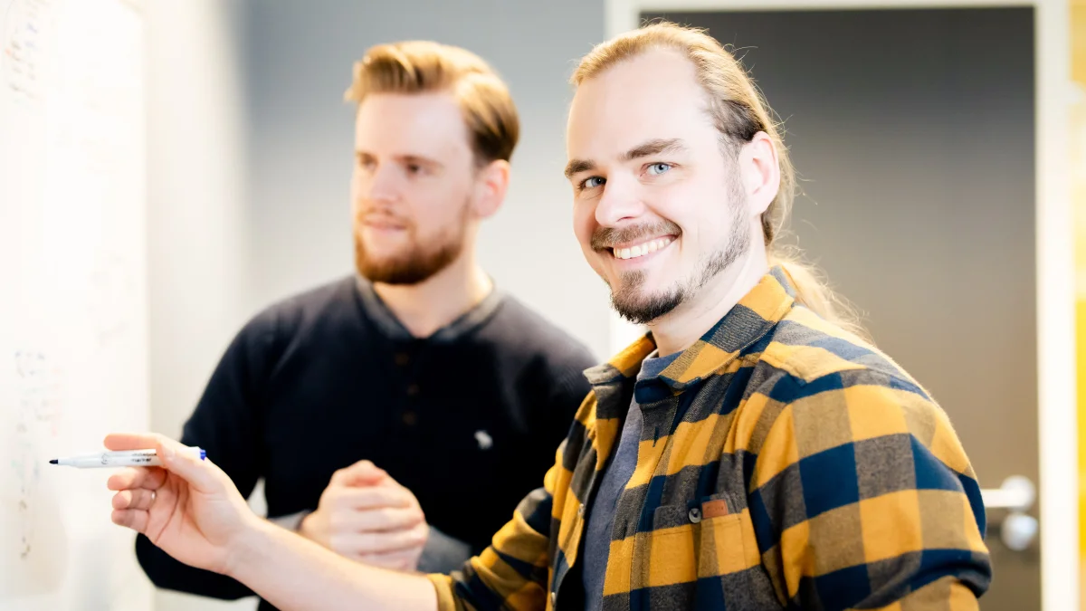 Breed lachende man die samen met een collega werkt aan een QHSE proces op een whiteboard.