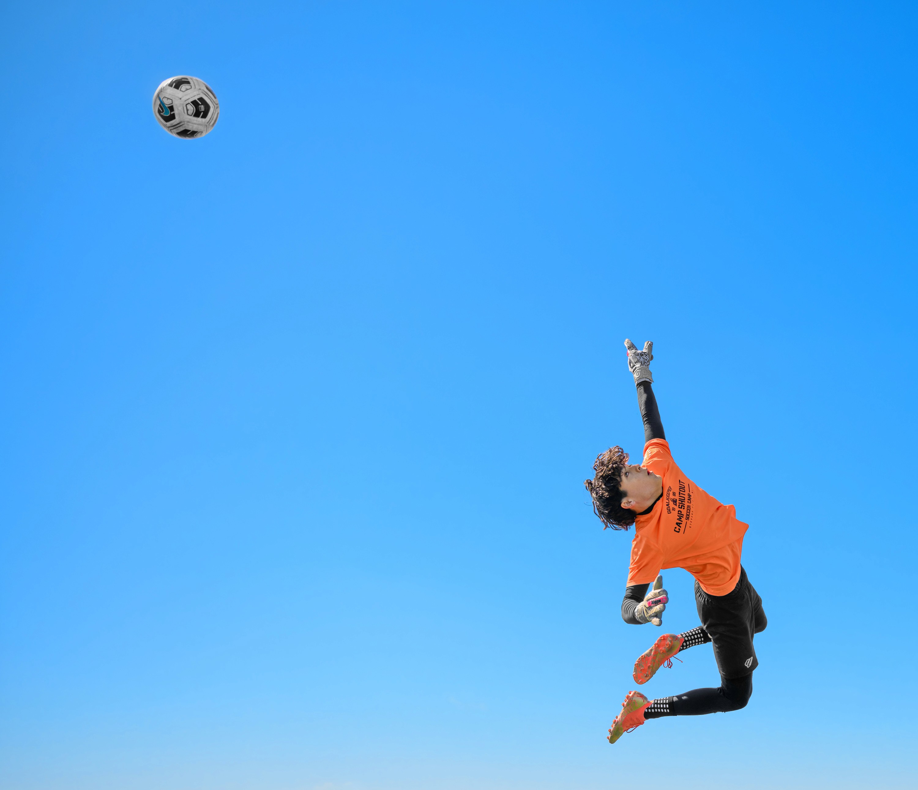 A youth goalkeeper in an orange jersey is suspended in midair against a bright blue sky, arching backward to watch the soccer ball after a deflection.