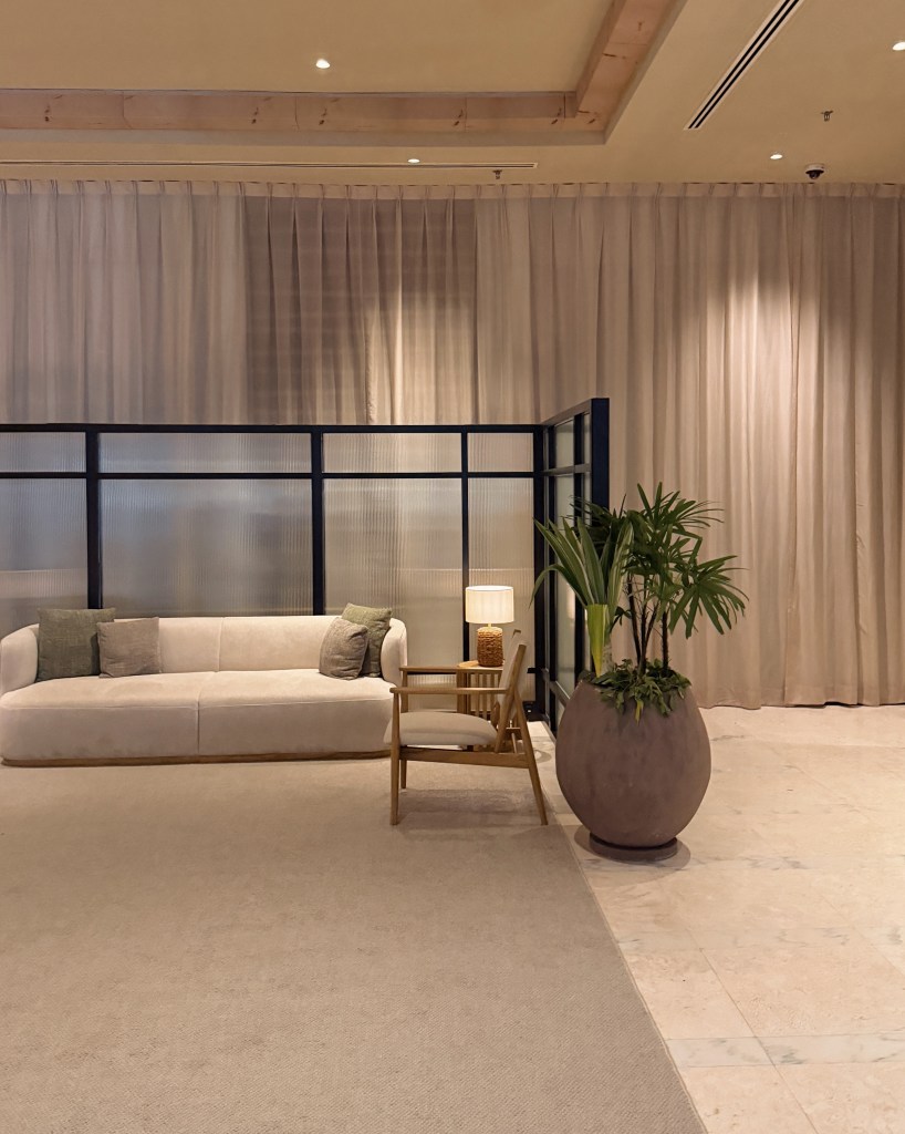 A stylish interior setting featuring a light-colored sofa with decorative pillows, a wooden chair, a potted plant, and a small lamp. The space is partially enclosed with a glass partition and has soft curtains in the background.