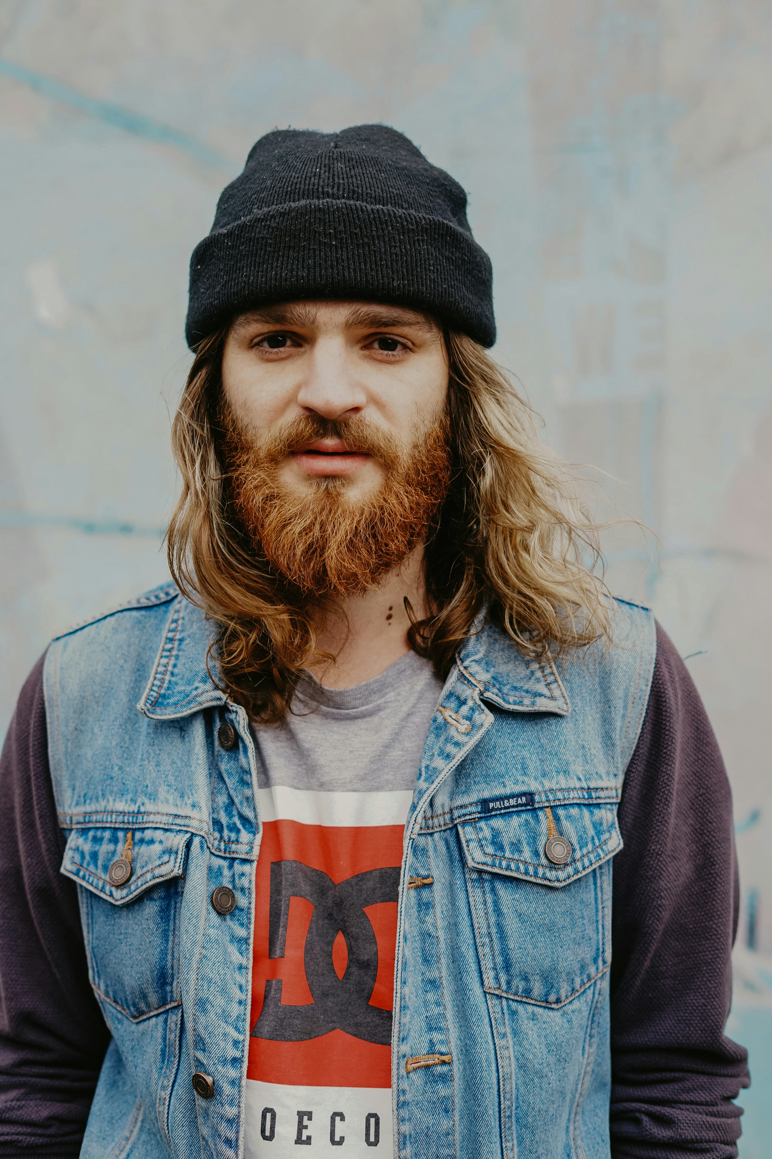 A person with long curly hair and a beard wears a beanie and denim vest against a neutral background.