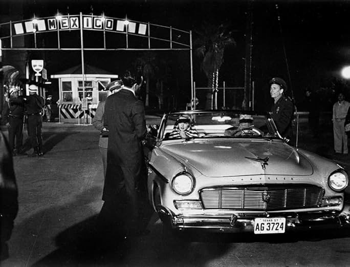 A vintage car parked in front of a theater entrance, with a person standing nearby in a nighttime scene.