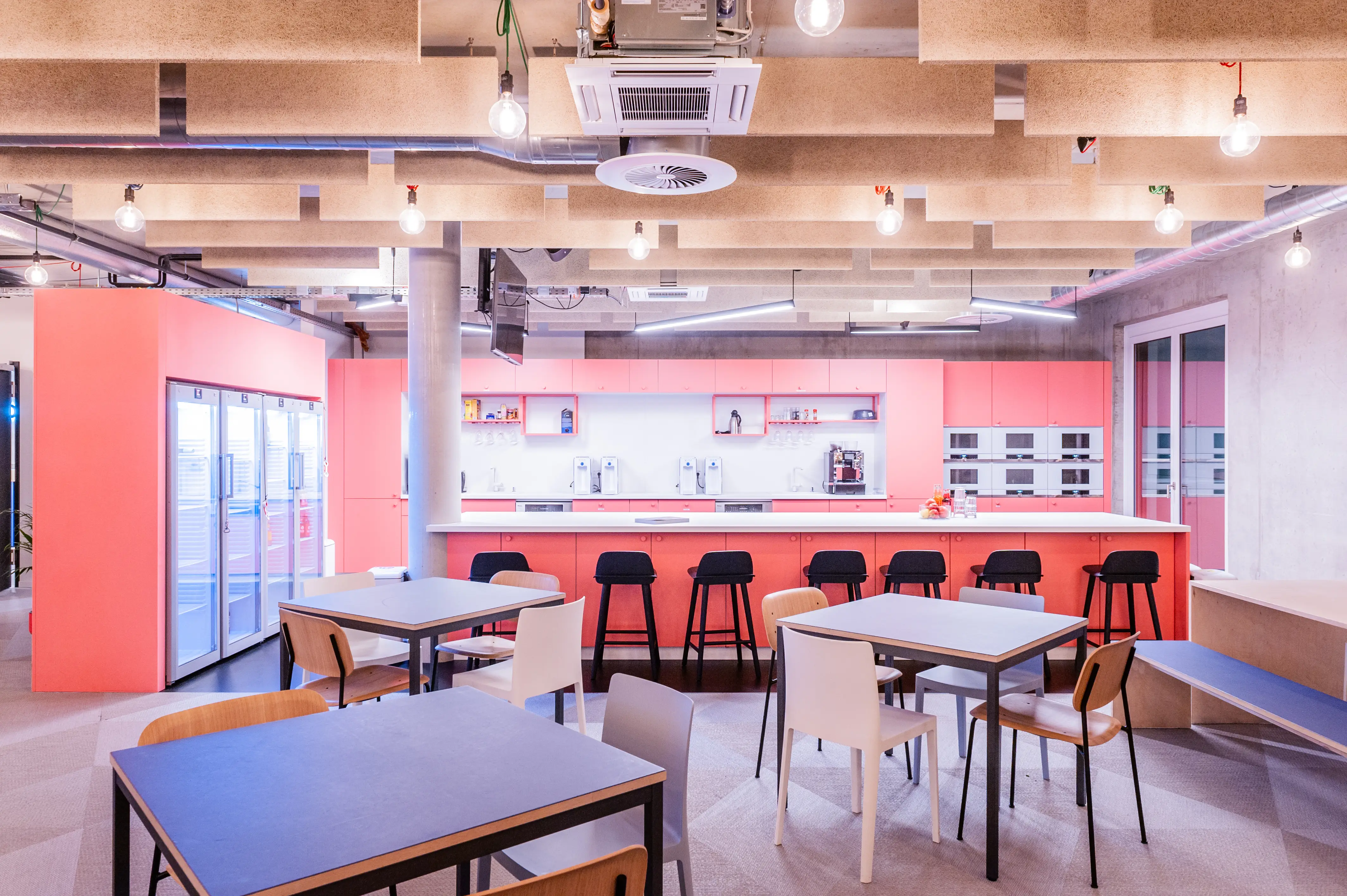 Office kitchen at OMIO with coral-pink cabinets, a long white island with black bar stools, glass-doored fridges, built-in ovens and tables with blue and white tops on grey carpet.