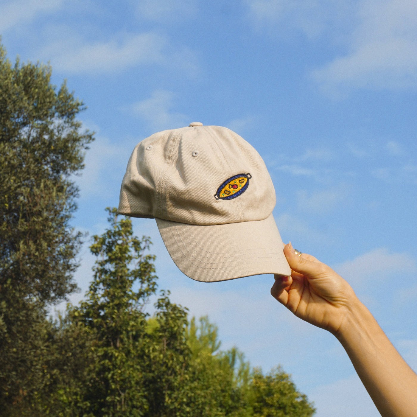 Beige cap with a paella design held against a blue sky with trees