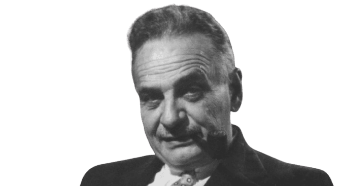 A black-and-white portrait of an older man with a pipe in his mouth, giving a contemplative expression. He has short, combed-back hair with a receding hairline, and is dressed in a suit jacket and collared shirt. The background includes shelves of books, suggesting an intellectual or scholarly setting.