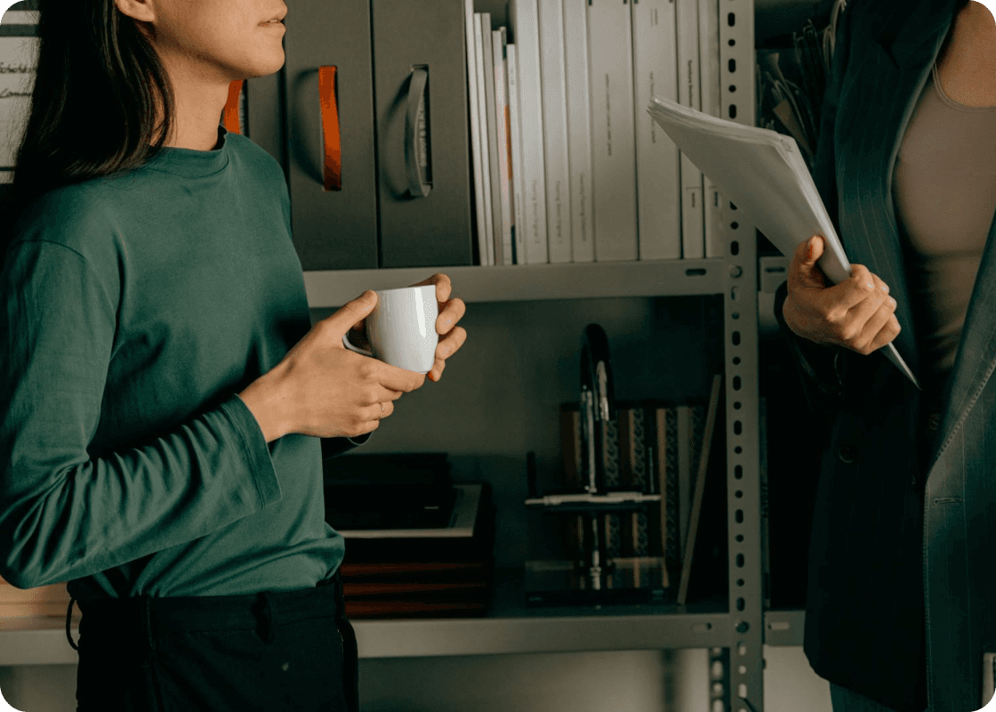 Two women stand in front of a desk, each holding a coffee cup, engaged in conversation