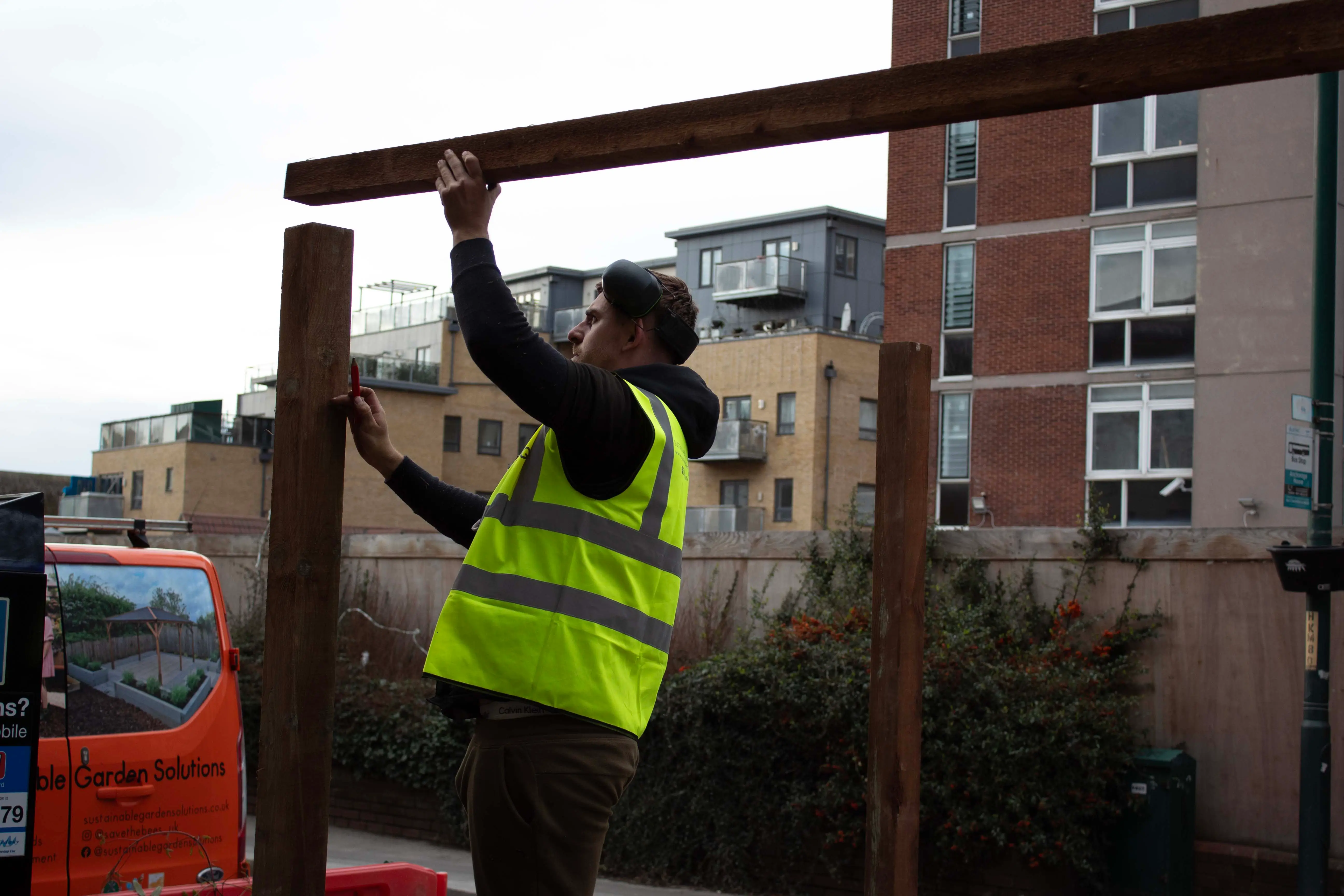 A worker in a yellow vest adjusts wooden beams at a construction site, with buildings in the background.