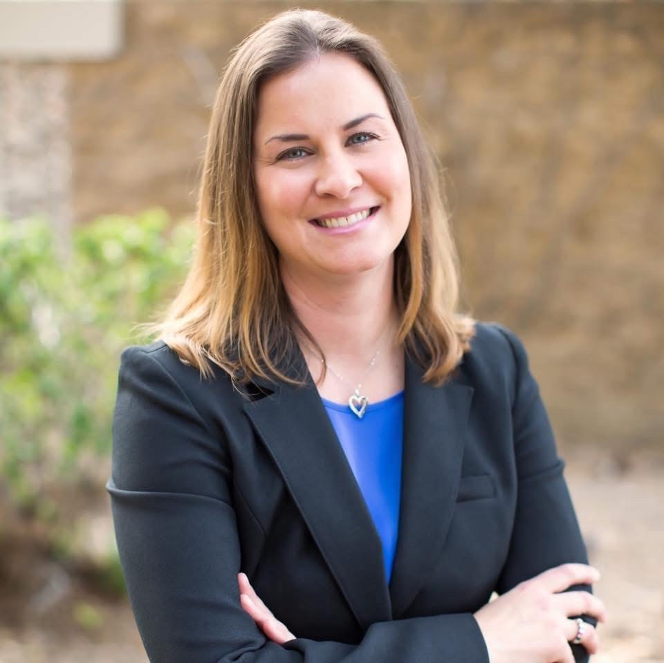 Image of a woman wearing a suit in a blue shirt and black coat with a tree in the background