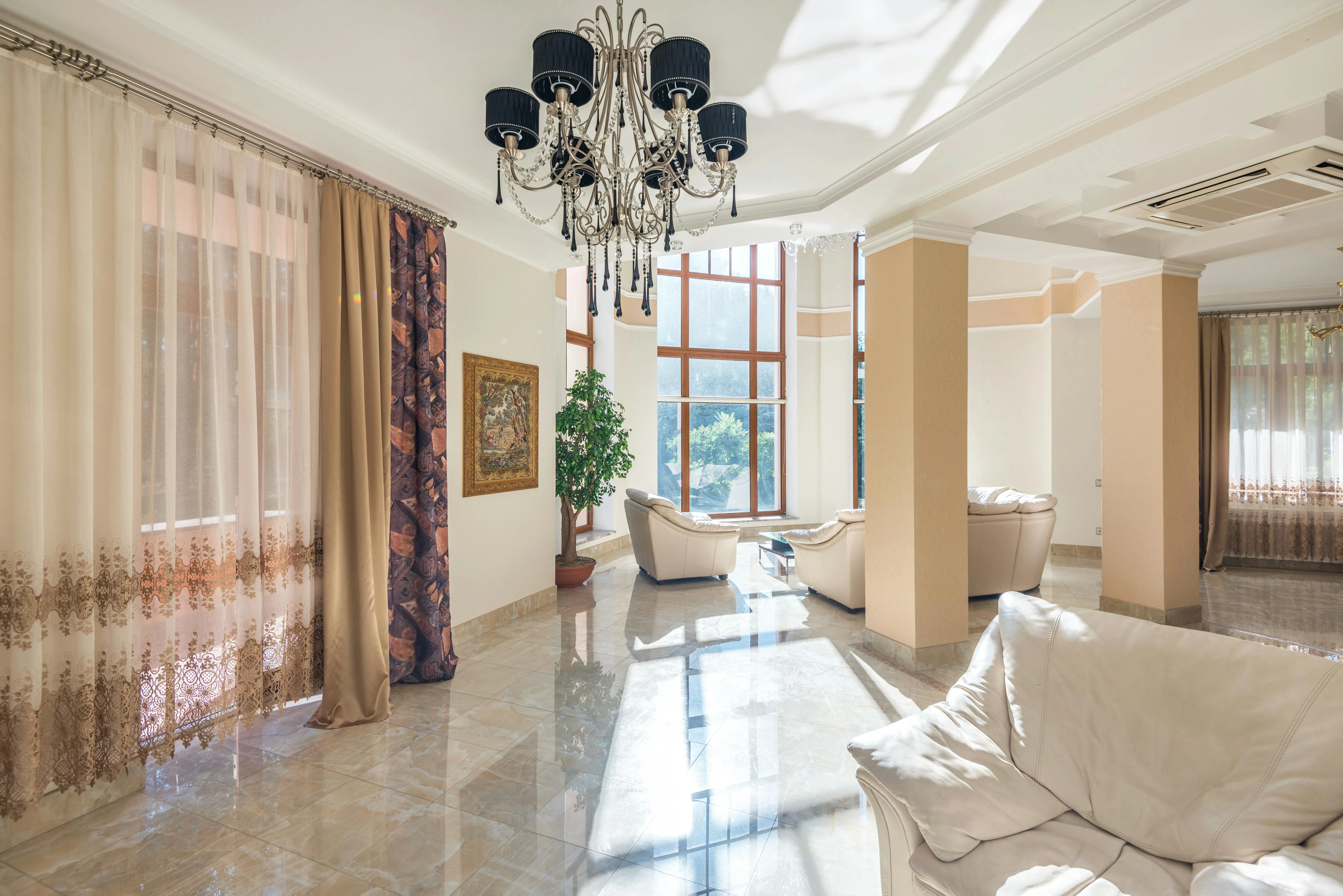 Bright, elegant living room with cream sofas and large windows. A black chandelier hangs from the ceiling, casting shadows on the marble floor.