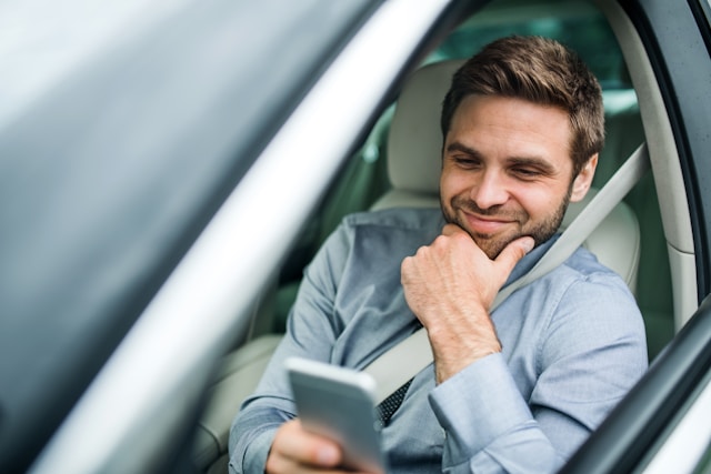 A man sitting in the driver’s seat of a car, smiling as he looks at his smartphone while resting his hand on his chin.