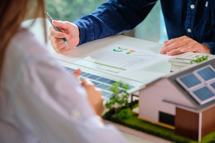 Zwei Personen besprechen Pläne an einem Tisch, wobei ein kleines Modellhaus und Dokumente zu sehen sind.