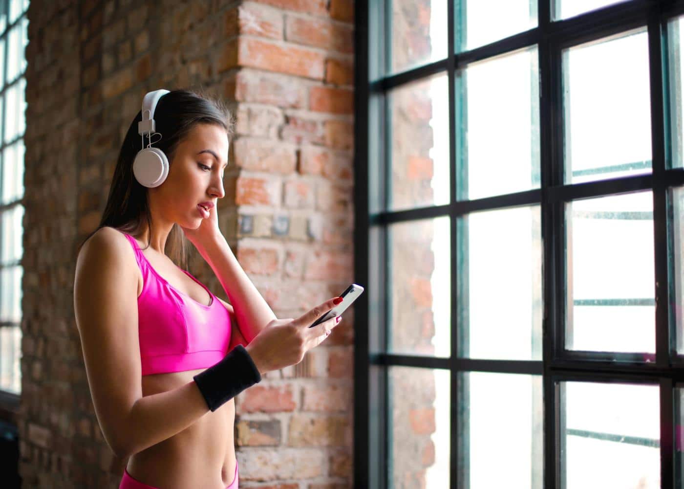 Woman indoors with headphones on and workout clothes looking at smartphone