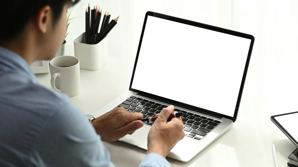 A person working on a laptop with a blank screen at a white desk, surrounded by office supplies and a coffee mug, focusing intently on the task at hand.