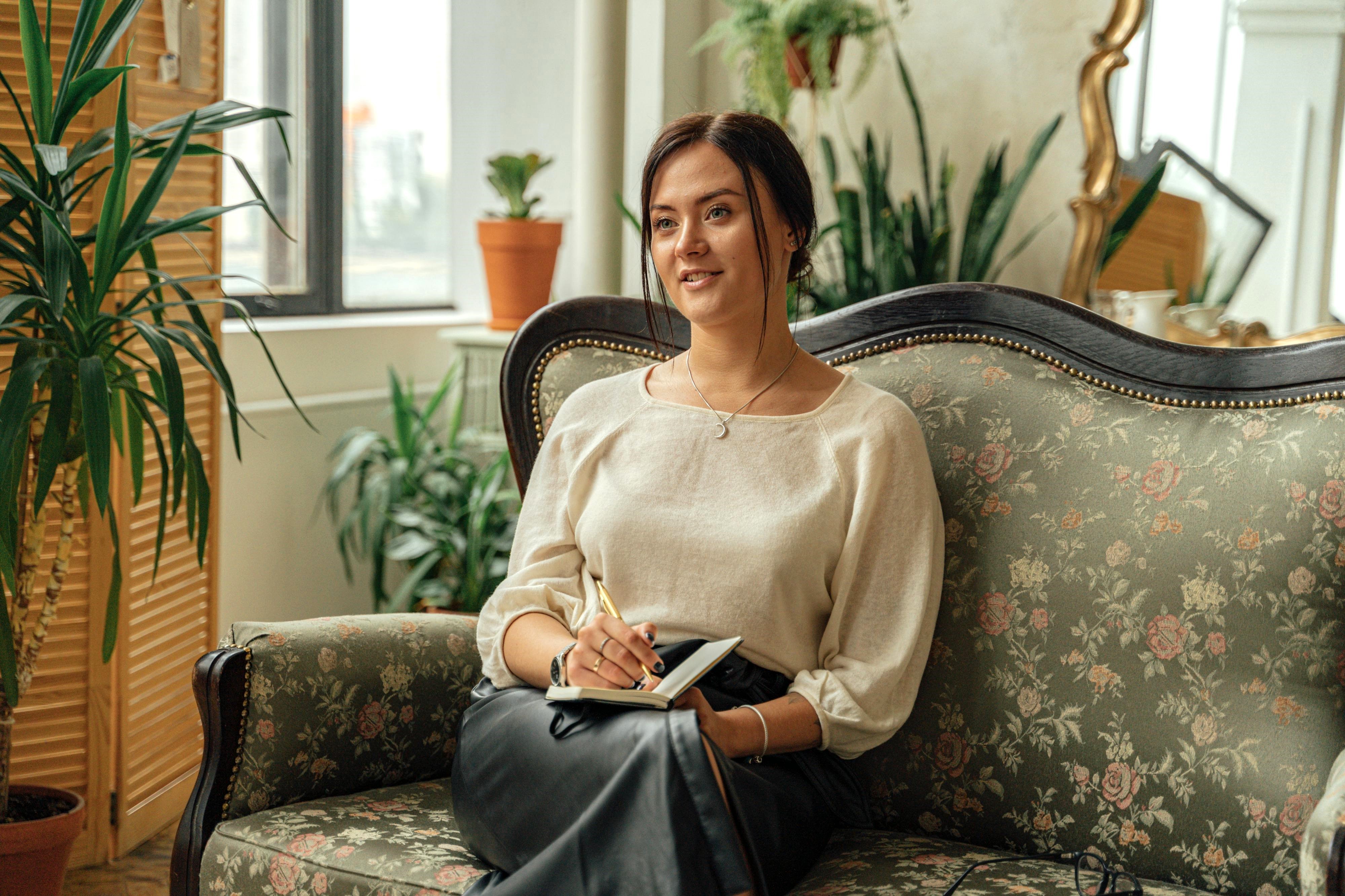 A therapist sits on a floral armchair holding a notebook and pen, attentively listening during a session.