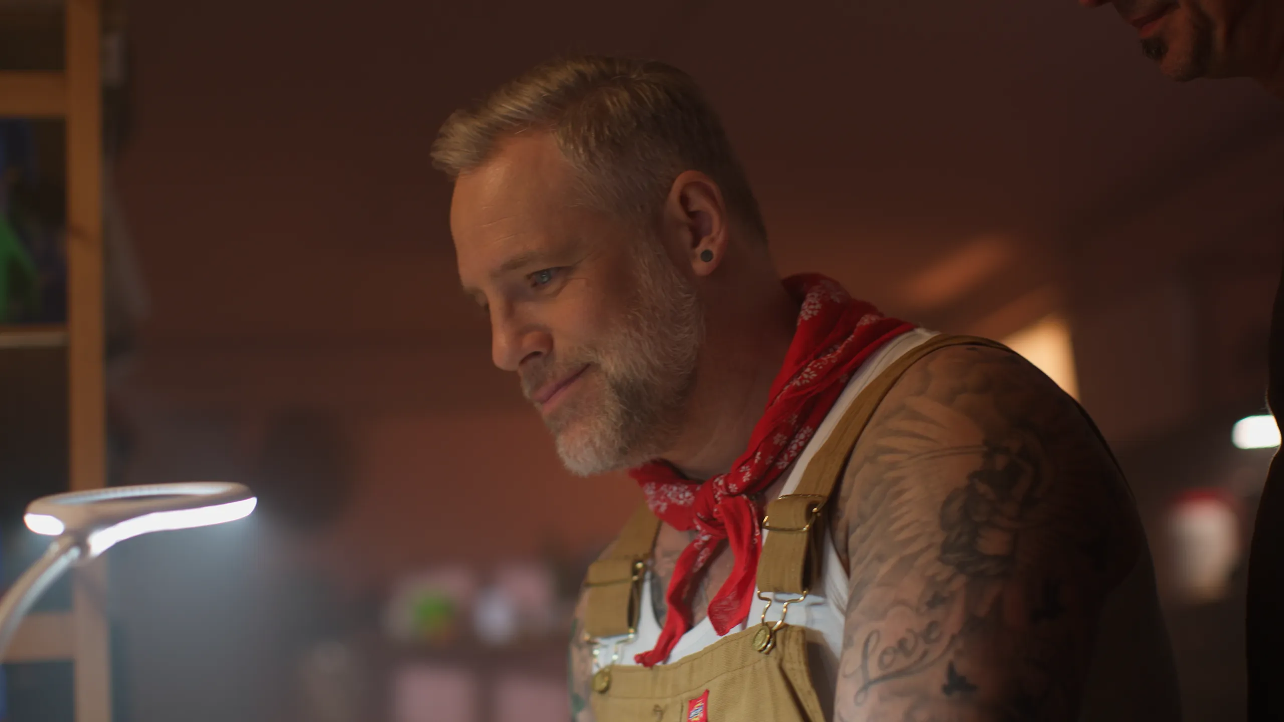 Close up portrait of a tattooed man in a red bandana and overalls looking down thoughtfully under a warm lamp.