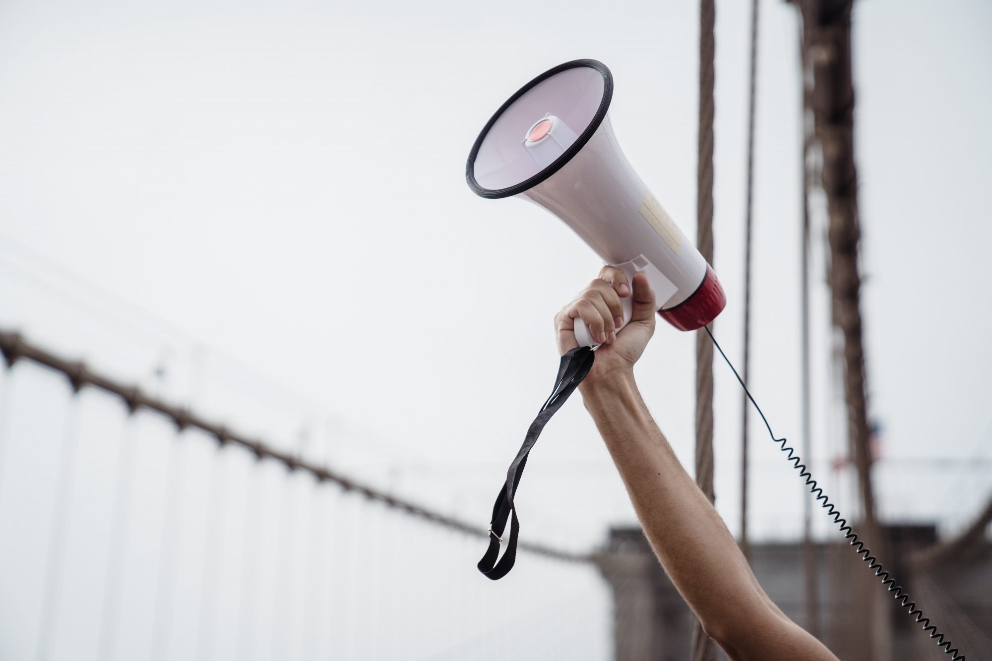 A hand holds a megaphone high, with a blurred background of cables and a cloudy sky.