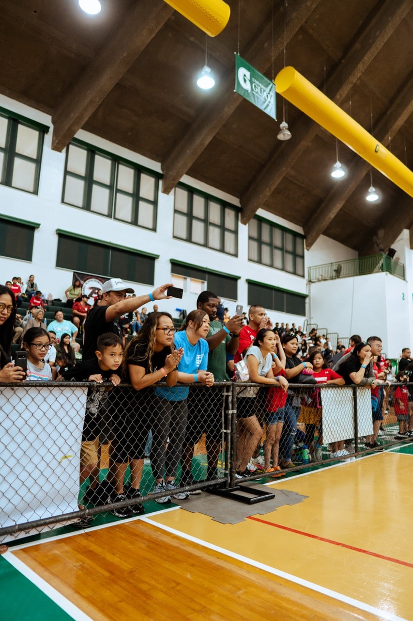 A crowd of enthusiastic spectators watches a jiu-jitsu match behind a chain-link fence at a Pacific Grappling Events tournament.