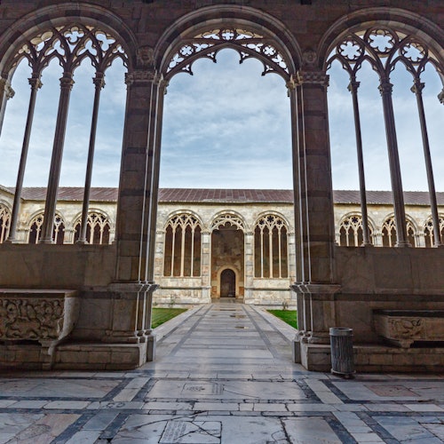 Cortile medievale con finestre ad arco in pietra, intricati intagli e un percorso piastrellato, incorniciato da colonne gotiche.
