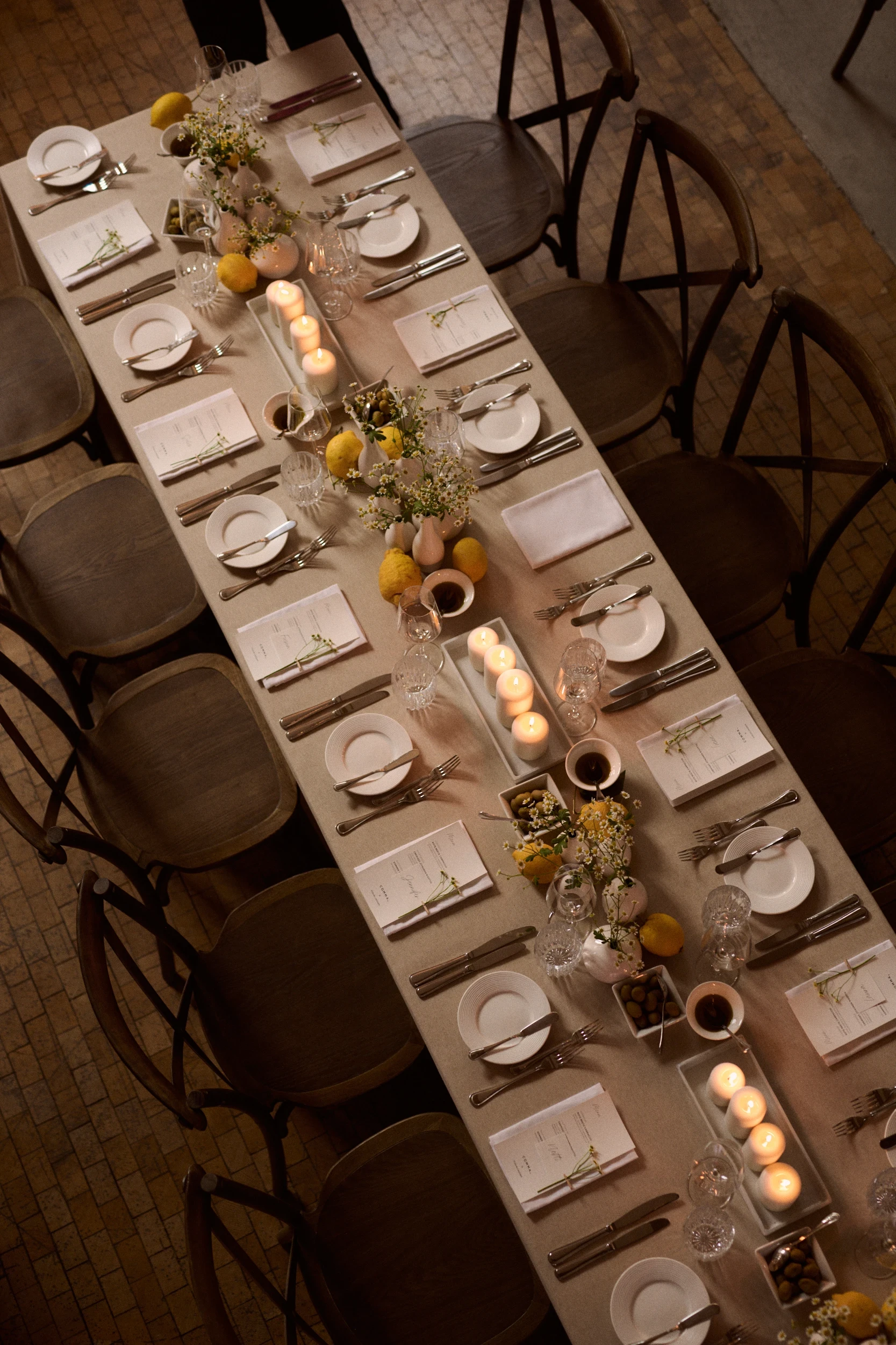Top view of the long dinner table with candles, lemons and menu cards between wooden chairs.