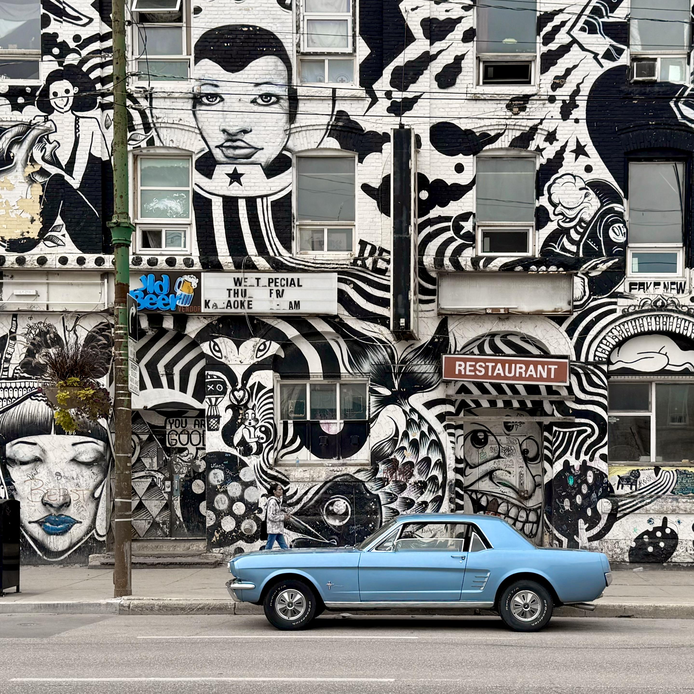 A building covered in black-and-white murals of faces and patterns, a pale blue vintage car parked along the street in front.