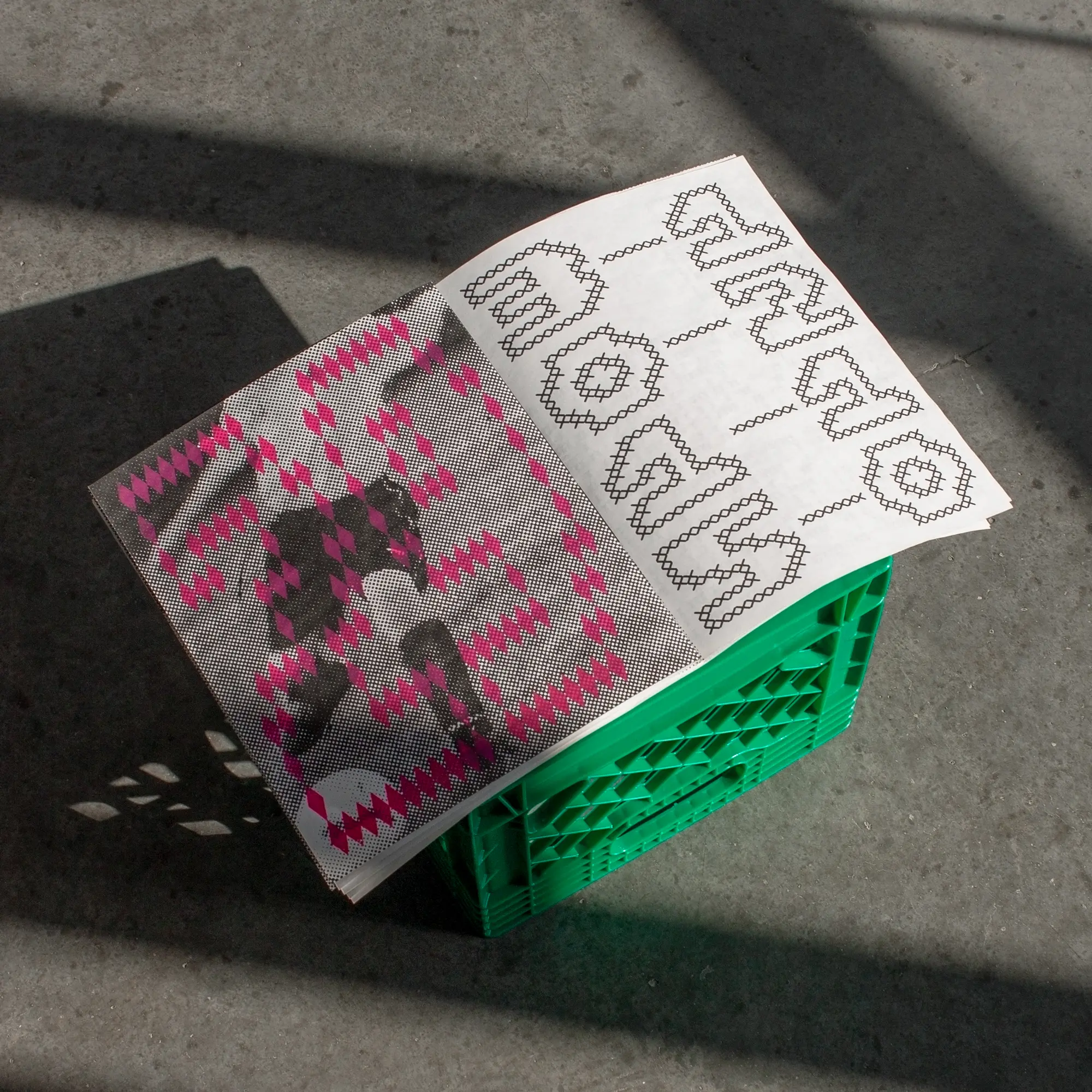 Journal ouvert posé sur une caisse verte, affichant des motifs typographiques expérimentaux en rose et noir sous une lumière naturelle.