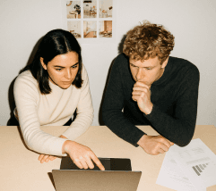 A young woman and man are focused on a laptop, discussing while sitting at a table. Papers are nearby.