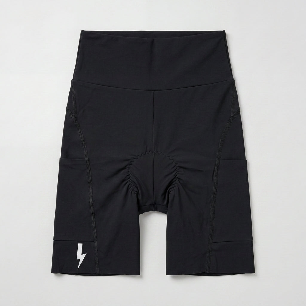 Flat-lay front view of LUCA® women's Empower padded cycling shorts in black. The design showcases a high waistband, integrated side pockets, ergonomic padding stitching at the crotch, and a signature white lightning bolt logo printed on the right bottom hem against a light grey background.- 