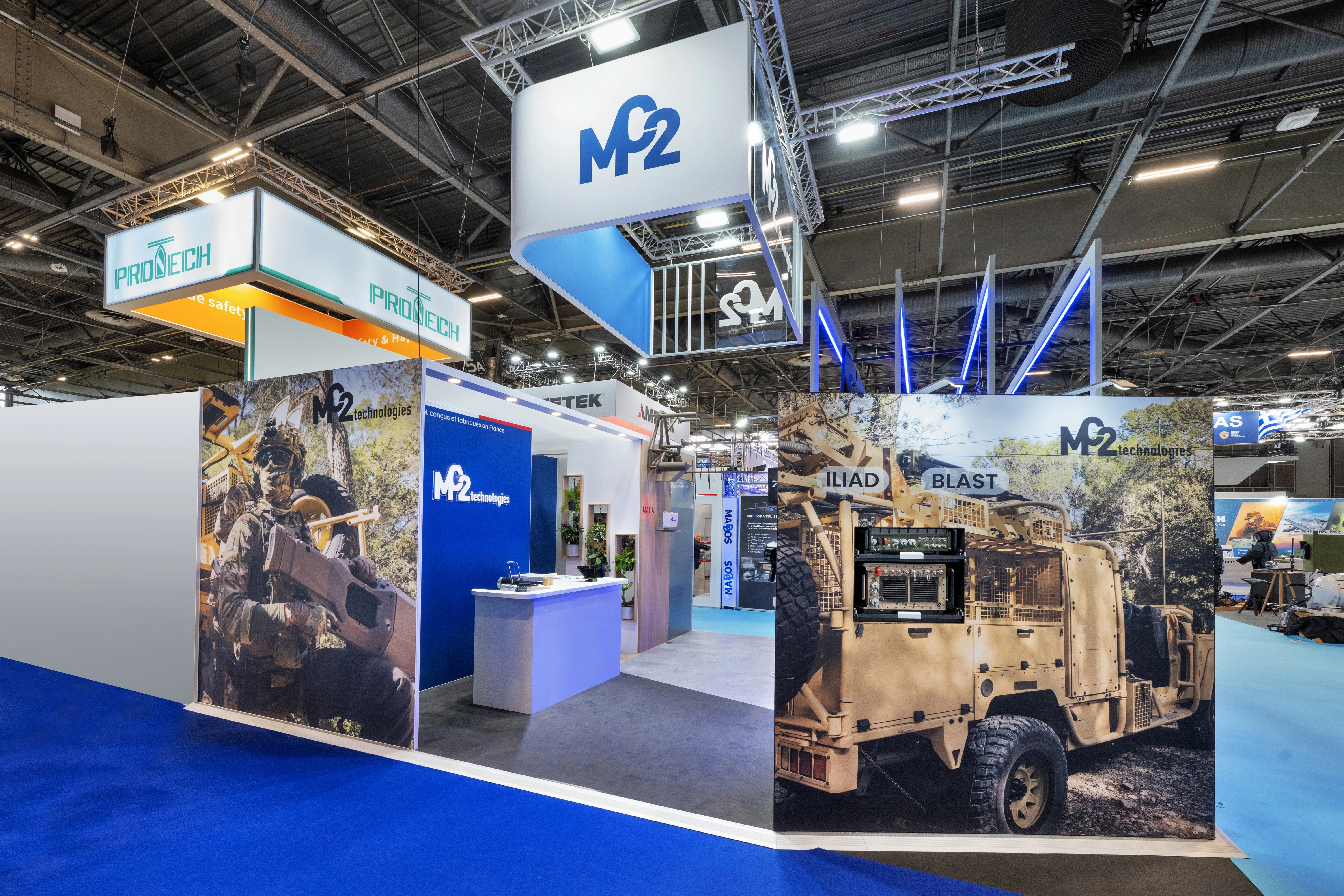 Stand MC2 Technologies lutte anti-drone conçu par EXPACE à Eurosatory 2026, Hall 5A — tour de brouilleurs MAJES DFB6 sur socle lumineux, mur jungle immersif et drones suspendus