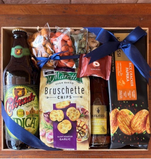 Cesta de presentes com cerveja artesanal, snacks, castanhas e geleia organizados em caixa de madeira decorada com laço azul.
