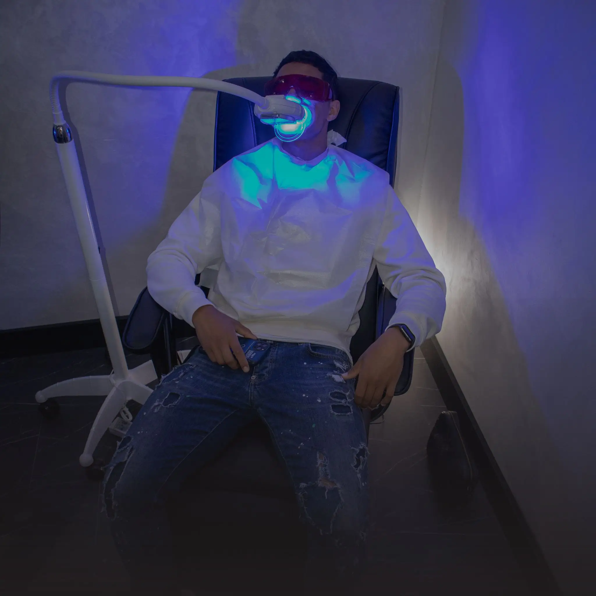 Een persoon zit in een stoel tijdens een tandverbleekbehandeling, met beschermende brillen en een witte schort, terwijl een blauw verlichte ruimte de atmosfeer creëert.