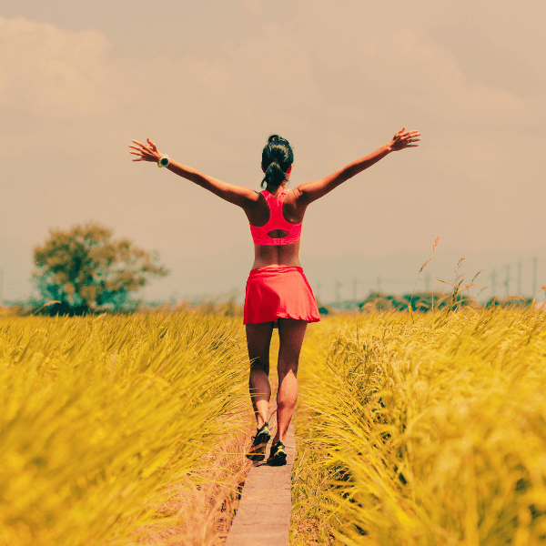 Woman walking in a field of yellow grass in pink workout wear with arms outreached to the sky - Red Light Therapy Bed Treatment at Live Supreme