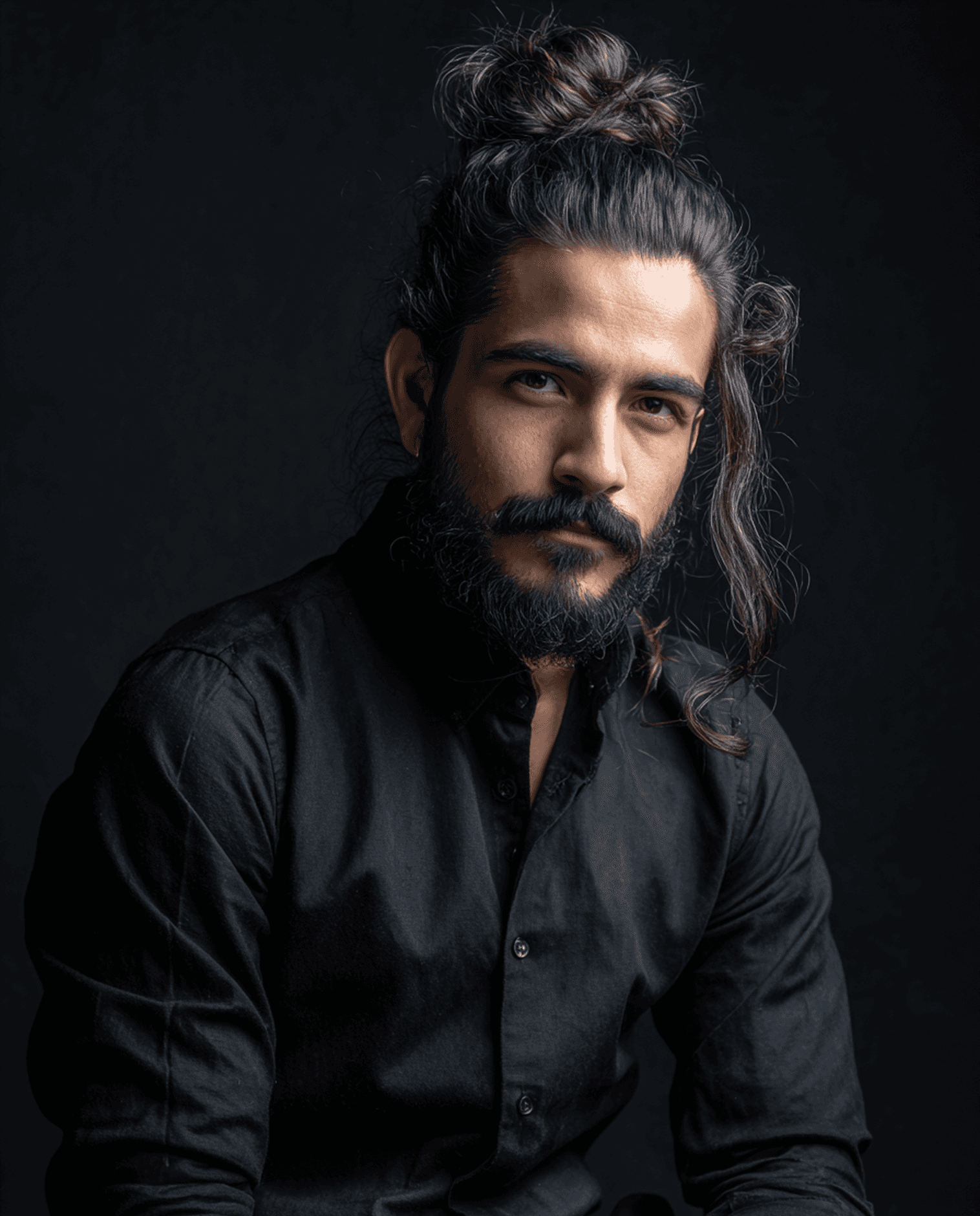 Man with a beard and tied-back hair against a dark background. He wears a black shirt and looks intently at the camera, conveying a thoughtful mood.