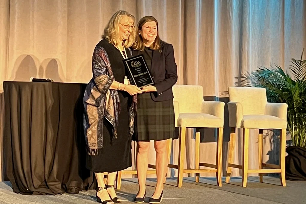 Two women smiling and standing together on stage, holding an award, with chairs and a backdrop in the background.