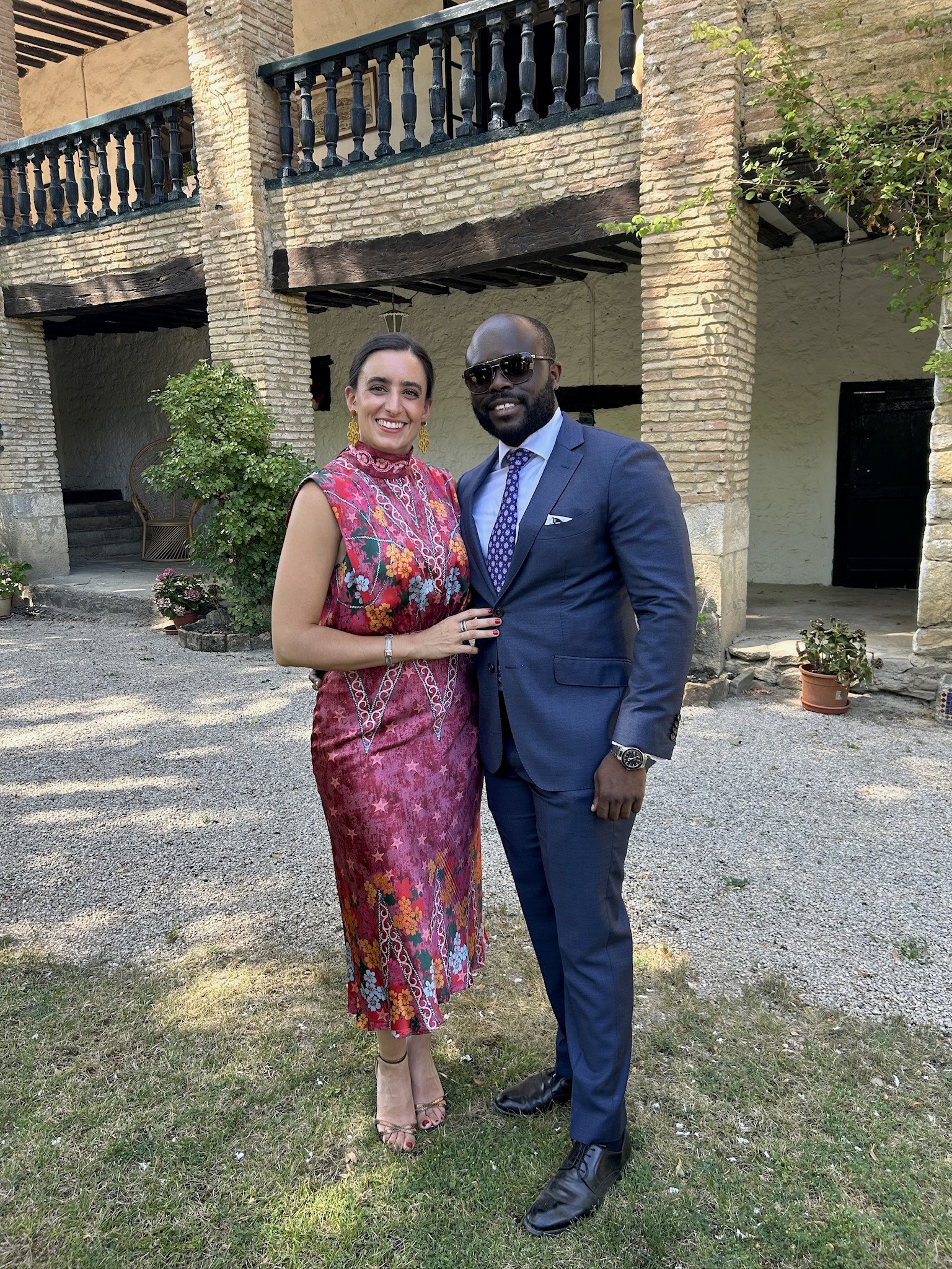 A groom in a dark suit carries a bride in a white dress across a sunny, grassy field. The couple shares a joyful, intimate moment with trees in the background.