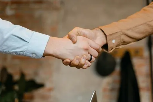 Close-up of two people shaking hands, symbolizing agreement or partnership. One wears a light blue shirt, the other a brown jacket.