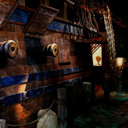 Wooden pirate ship with cannons, ropes, netting, and a lantern, docked at a pier with posts and ropes in dim lighting.