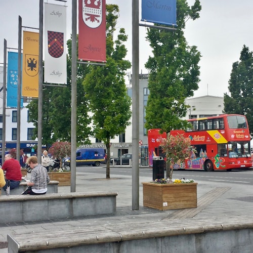 אנשים יושבים ליד כיכר עם תרנים, עצים ואוטובוס דו-קומתי אדום ברקע ביום מעונן.