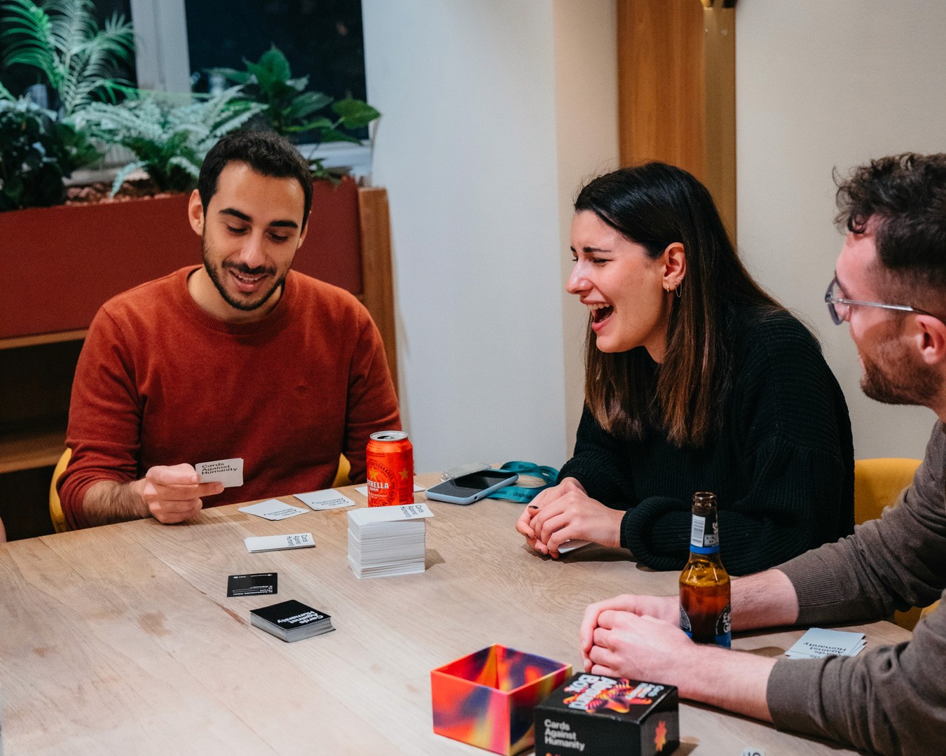 Three friends are seated around a table, enjoying a game and sharing laughs in a cozy indoor setting.