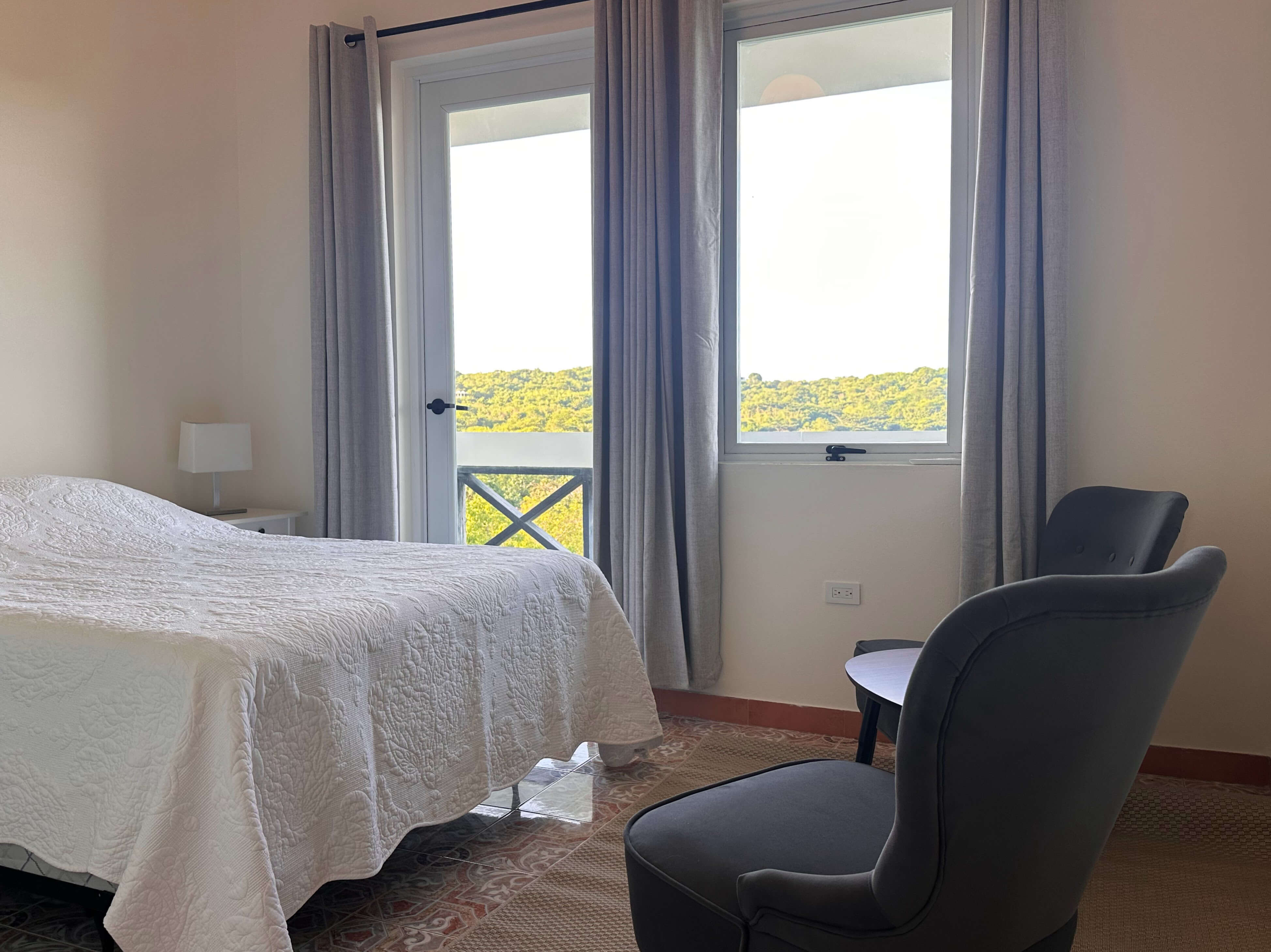 Queen bed with two side tables and lamps in Room 8 at Club Vieques, with a view of the surrounding tropical landscape through large windows.