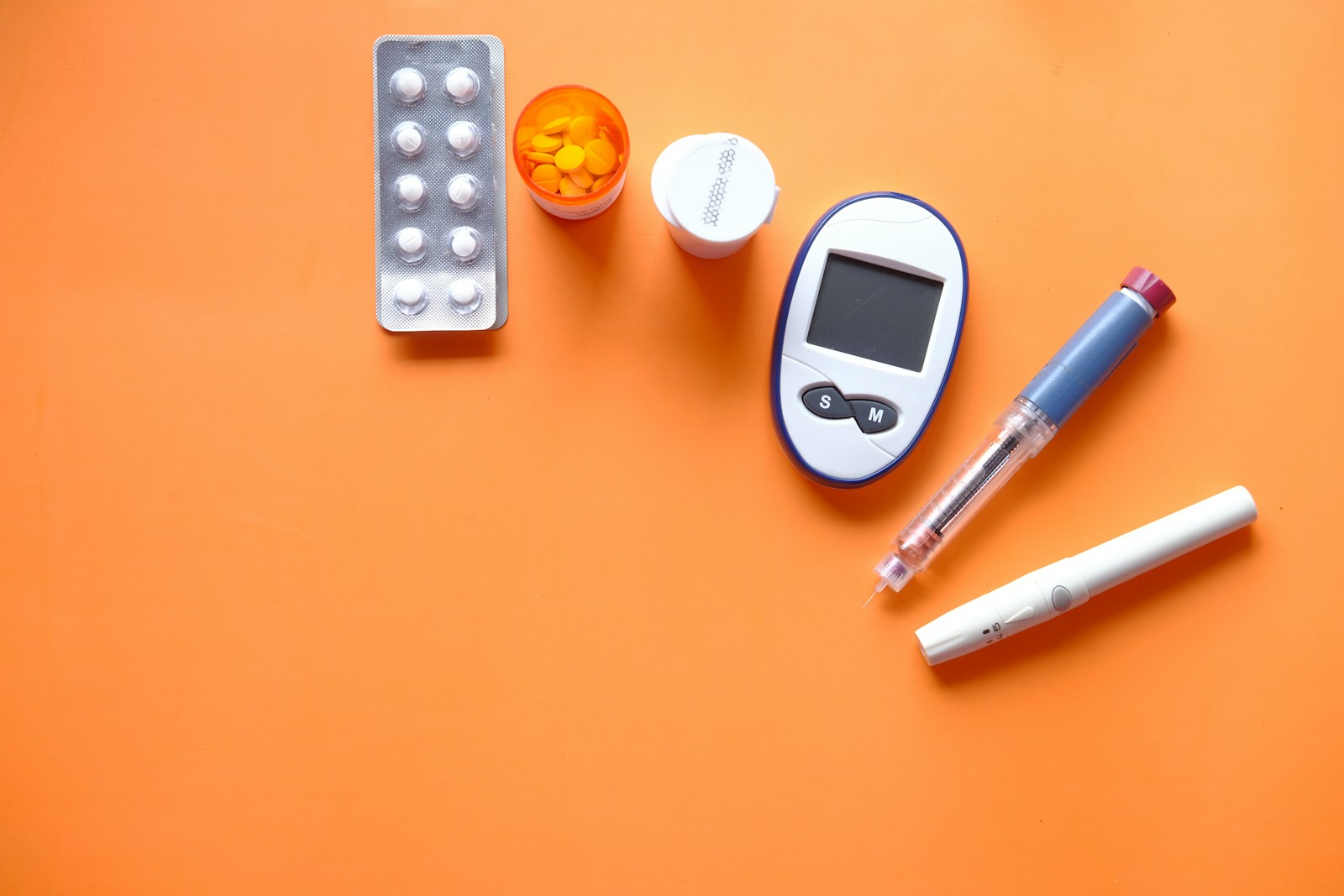 A blood glucose meter, insulin pen, lancet, and medication laid out on an orange surface, representing diabetes management