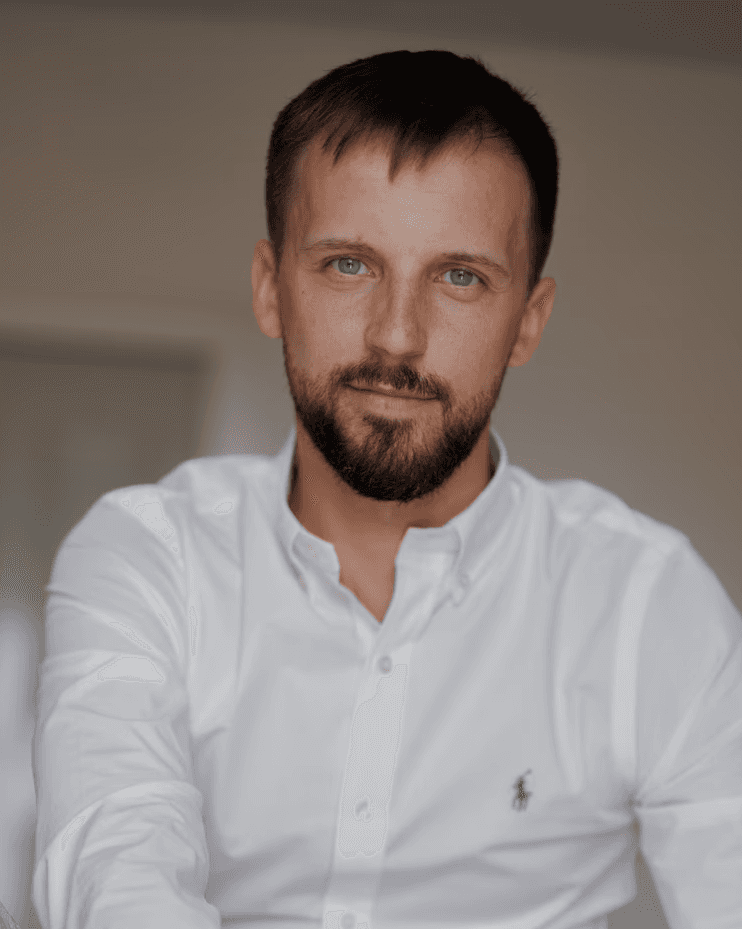 Close-up portrait of a person wearing a gray insulated hooded jacket with the hood up, showing their face framed by the hood against a light background.