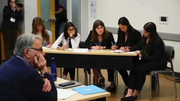 High school students participating in a debate competition, taking notes and engaging in critical thinking during a judged academic event for high schoolers.