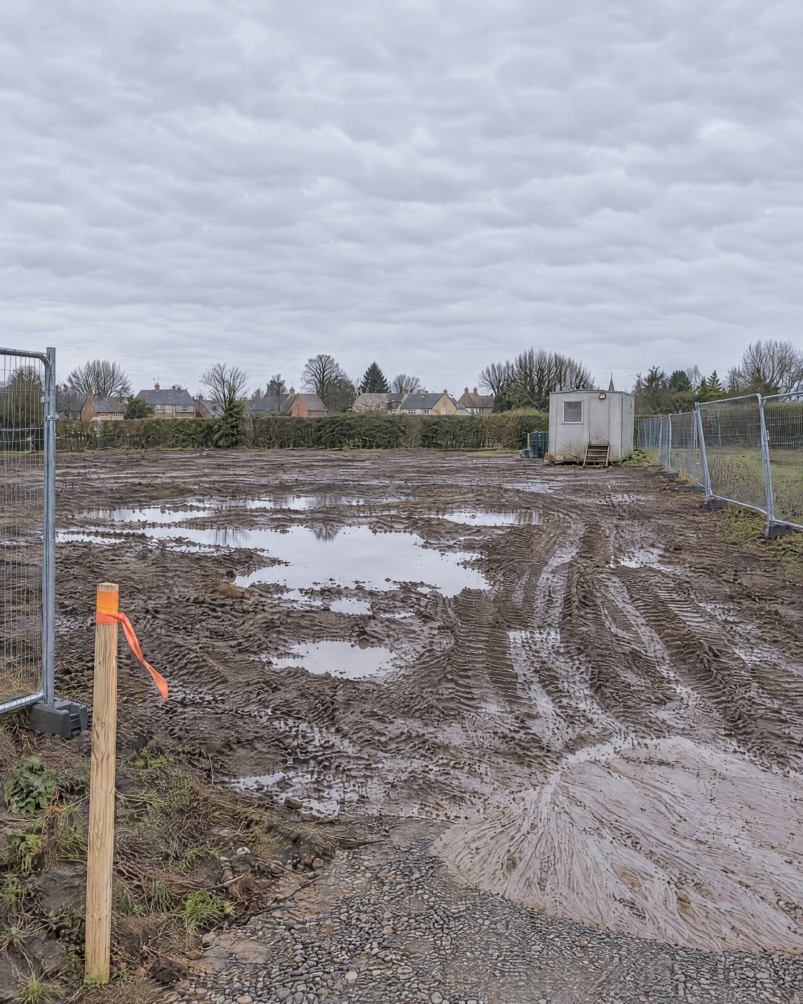 The cleared plot from the gateway, tyre tracks in wet mud and houses beyond the hedgerow