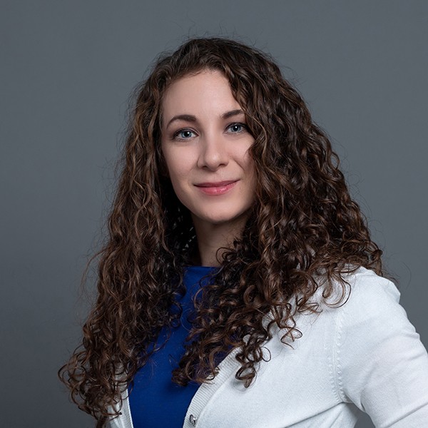 A photo of Rachael, a woman with fair skin and long brown curly hair. She wears a white cardigan and blue dress