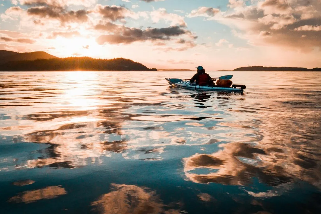 Person in a kayak on the water at sunset.  Pink clouds above, and glassy water.