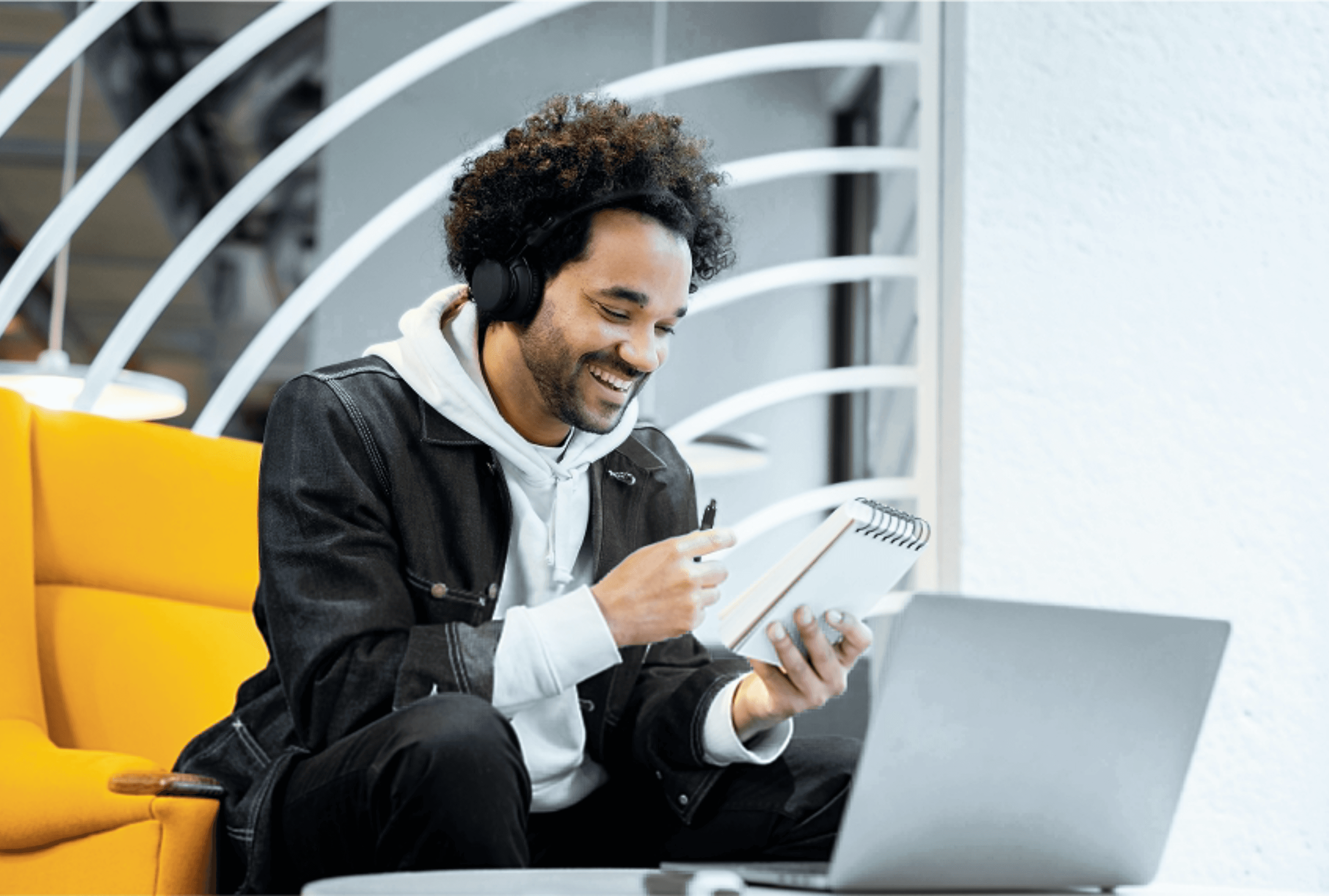 A smiling man with curly hair sitting on a yellow chair, holding documents and working on a laptop, representing productivity and a positive work environment.