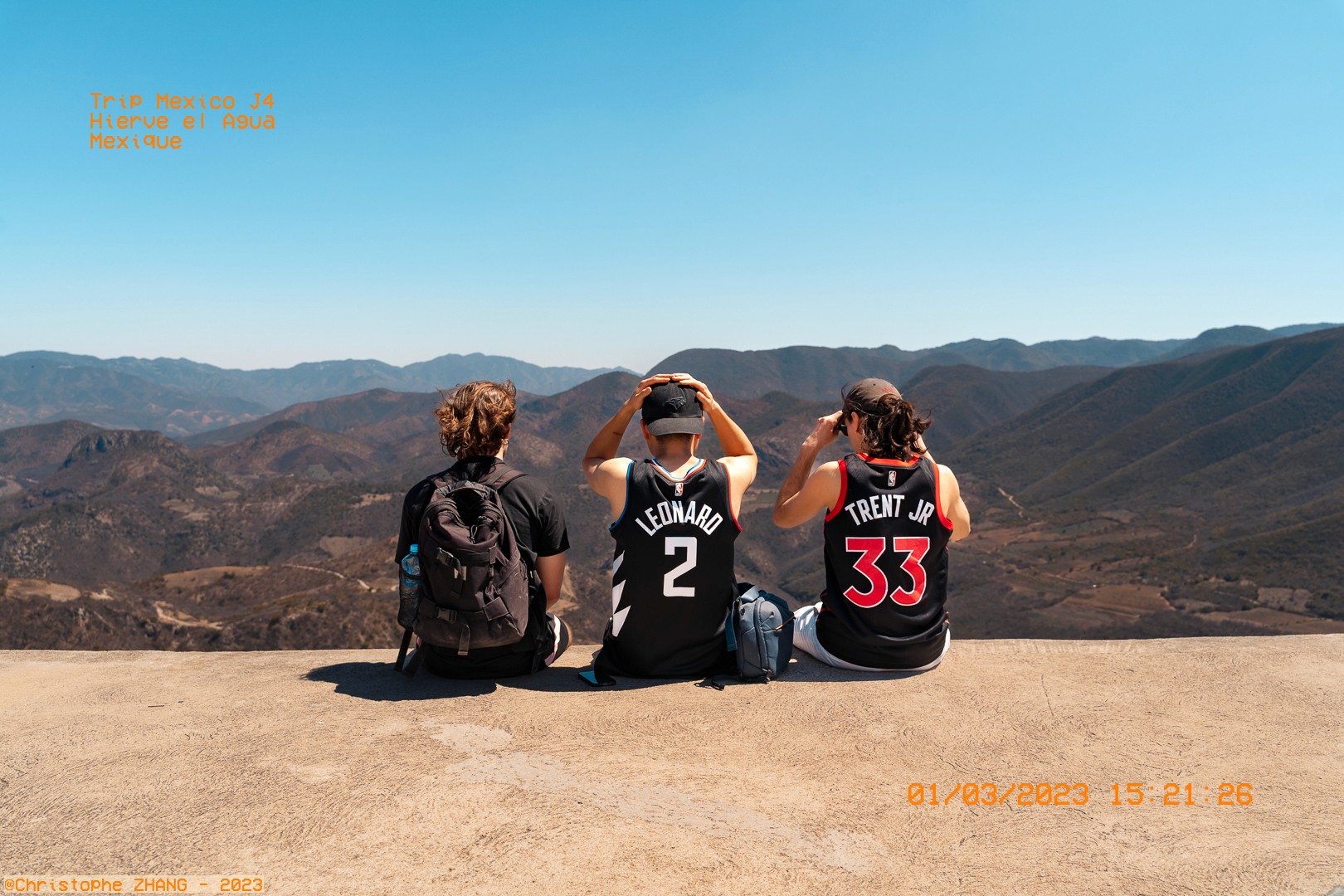Trois amis assis face aux montagnes de Hierve el Agua, portant des maillots de basket sous un ciel dégagé.