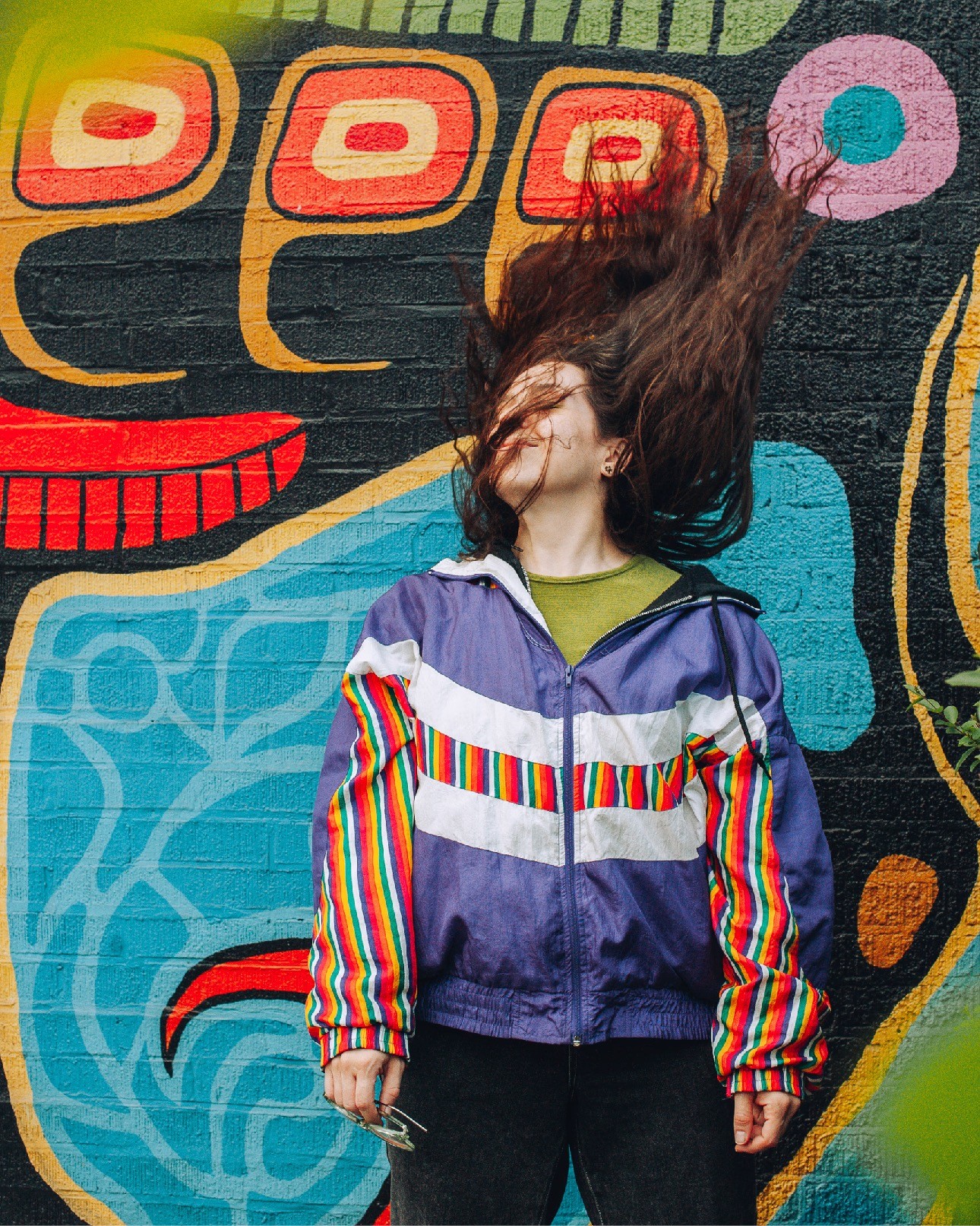 Person wearing a colourful patterned jacket standing in front of a bold Indigenous-style mural