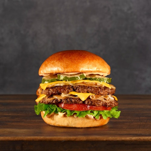 深色背景下的木质台面上放着一个双层芝士汉堡,配有生菜、番茄、酸黄瓜和酱料。