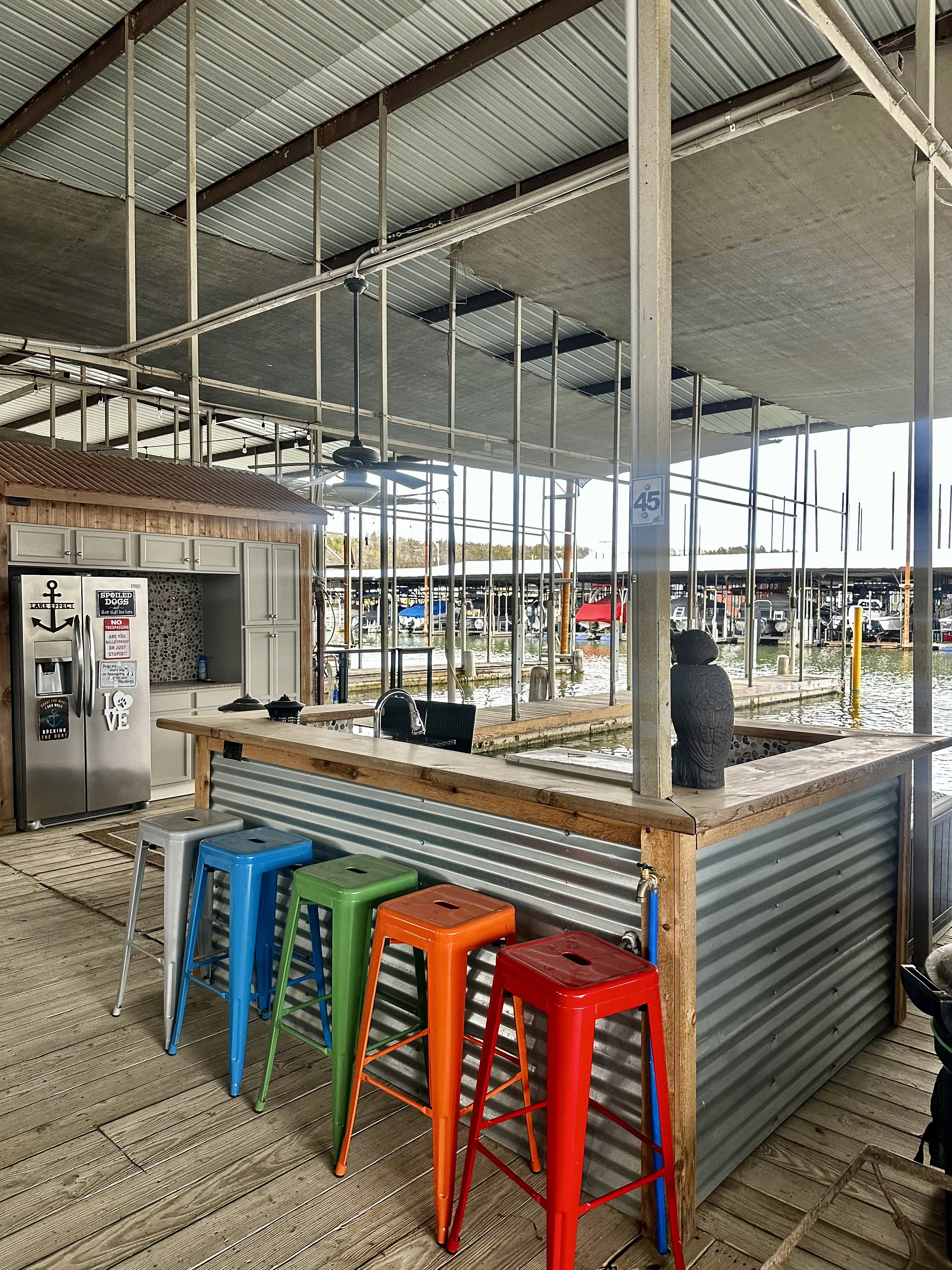 A vibrant waterfront bar area features a corrugated metal counter with colorful bar stools in red, orange, green, and blue, set under a corrugated roof with a scenic view of a marina filled with boats in the background.